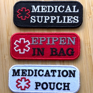May include: Three rectangular patches with rounded corners. The top patch is black with white text reading "MEDICAL SUPPLIES" and a red medical symbol. The middle patch is red with gray text reading "EPIPEN IN BAG" and a white medical symbol. The bottom patch is light blue with black text reading "MEDICATION POUCH" and a red medical symbol.
