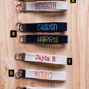May include: Seven fabric wristlet keychains with metal hardware and embroidered names. The text FONT STYLE OPTIONS is visible. The names include geni, aniyah, cayden, harris, jayla b., kayla, and london in various thread colors and fonts.