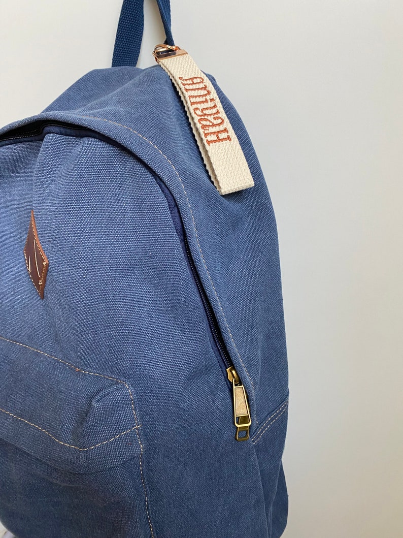 May include: A blue canvas backpack featuring a brown diamond-shaped patch on the front pocket and a cream-colored fabric zipper pull tag embroidered with the name amiyah in brown thread, complete with a dark blue top handle and metal zipper.