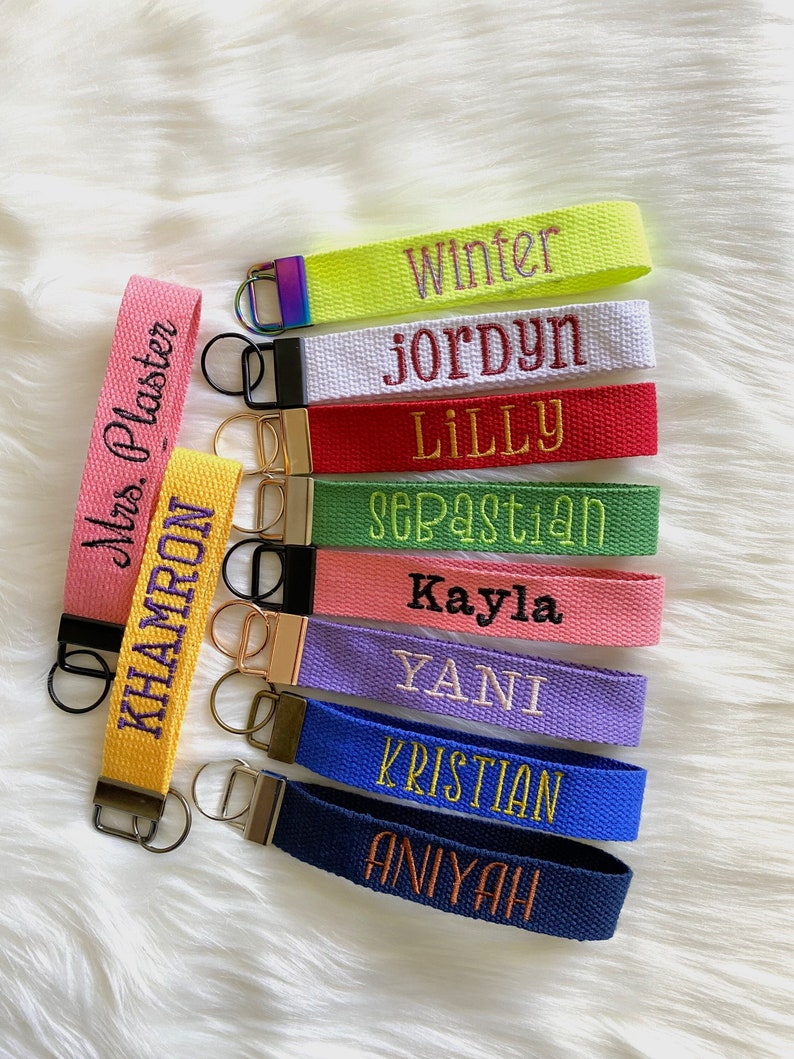 May include: A collection of ten fabric wristlet keychains in various colors including neon yellow, white, red, green, pink, purple, blue, and navy, each featuring embroidered names like Winter, Jordyn, Lilly, Sebastian, Kayla, Yani, Kristian, Aniyah, Mrs. Plaster, and Khamron.