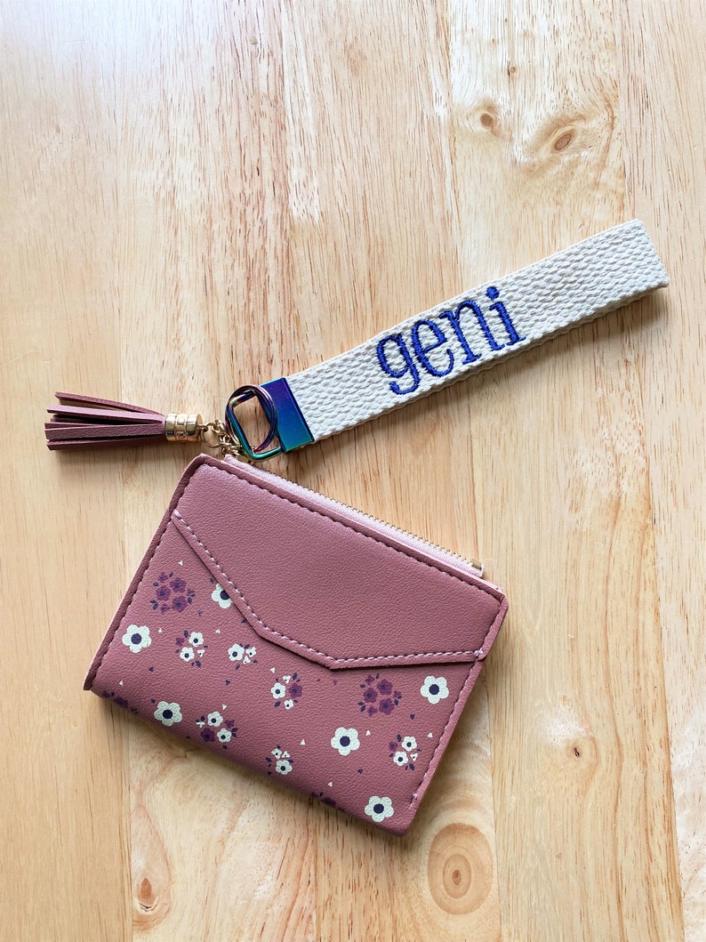 May include: A mauve wallet with a small floral pattern, featuring a zipper closure and a matching mauve tassel. Attached is a beige fabric wristlet strap embroidered with the name "geni" in blue thread, connected by a rainbow-colored metal ring.