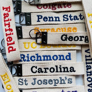 May include: A collection of off-white fabric keychains featuring embroidered university names including Temple, Tampa, Colgate, Penn State, Syracuse, Georgia, UC Boulder, Richmond, Carolina, St Joseph's, Scranton, Elon, Villanova, and UVM.