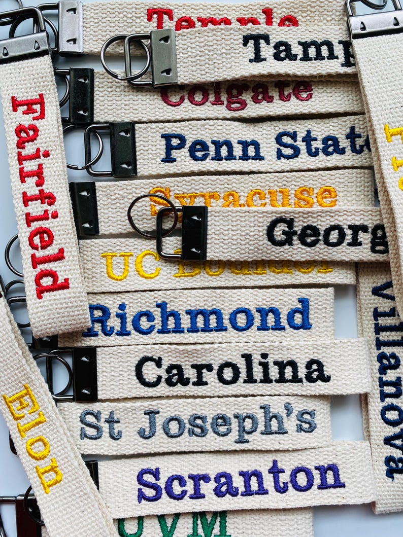 May include: A collection of off-white fabric keychains featuring embroidered university names including Temple, Tampa, Colgate, Penn State, Syracuse, Georgia, UC Boulder, Richmond, Carolina, St Joseph's, Scranton, Elon, Villanova, and UVM.