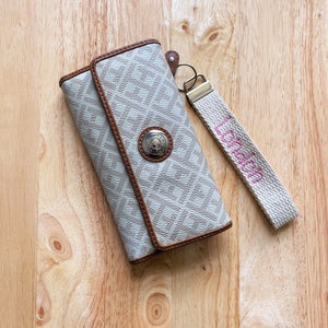 May include: A beige and brown patterned wallet with a silver-toned circular snap closure featuring the Tommy Hilfiger logo. Attached to the wallet is a beige fabric wrist strap with the word London embroidered in pink thread.
