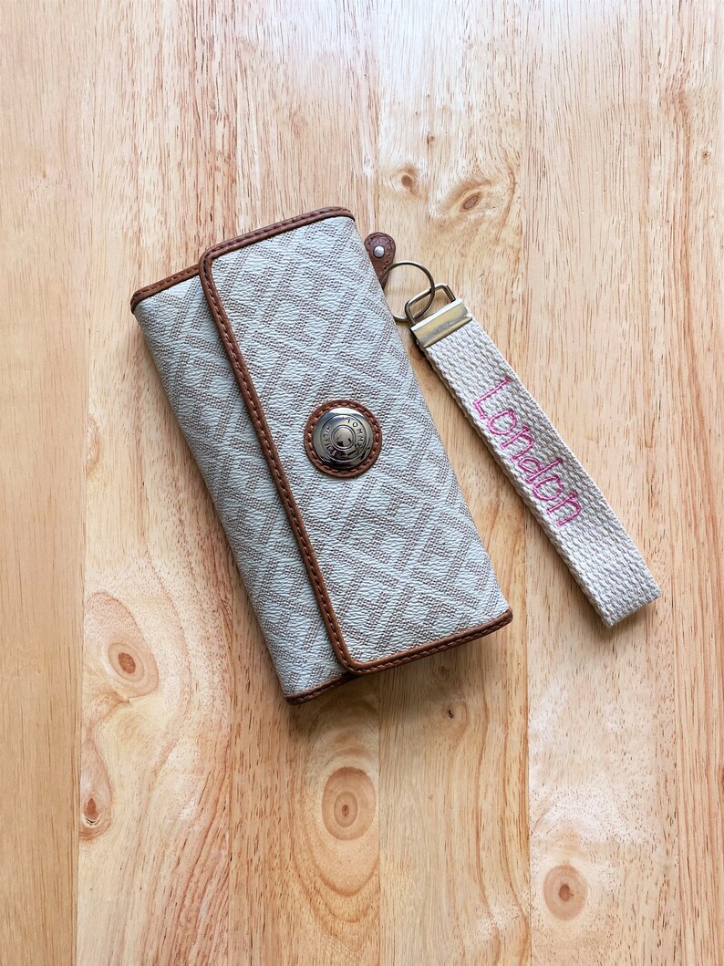 May include: A beige and brown patterned wallet with a silver-toned circular snap closure featuring the Tommy Hilfiger logo. Attached to the wallet is a beige fabric wrist strap with the word London embroidered in pink thread.