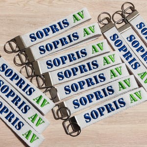 May include: A collection of white fabric keychains featuring the embroidered text SOPRIS AV in dark blue and bright green thread. Each keychain is attached to a silver-toned metal split ring and a rectangular metal clamp at the top end.