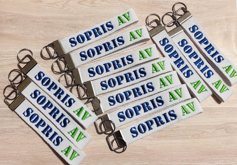 May include: A collection of white fabric keychains featuring the embroidered text SOPRIS AV in dark blue and bright green thread. Each keychain is attached to a silver-toned metal split ring and a rectangular metal clamp at the top end.