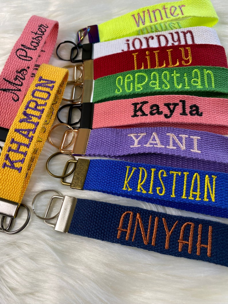 May include: A collection of fabric wristlet keychains in various colors including neon yellow, white, red, green, pink, purple, blue, navy, and yellow, each featuring embroidered names like Winter, Jordyn, Lilly, Sebastian, Kayla, Yani, Kristian, Aniyah, Mrs. Plaster, and Khamron with metal hardware.
