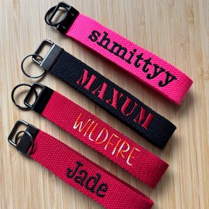 May include: Four fabric keychains with metal rings. The top is hot pink with black text reading shmittyy. The second is black with red text reading MAXUM. The third is red with orange and yellow text reading WILDFIRE. The bottom is red with black text reading Jade.