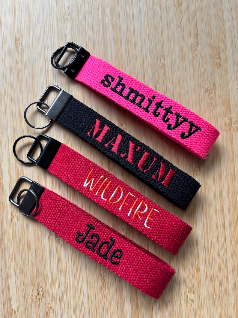 May include: Four fabric keychains with metal rings. The top is hot pink with black text reading shmittyy. The second is black with red text reading MAXUM. The third is red with orange and yellow text reading WILDFIRE. The bottom is red with black text reading Jade.