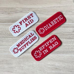 May include: Four rectangular embroidered patches with rounded corners. Two are white with red text and a red medical symbol: "FIRST AID" and "MEDICAL SUPPLIES". The other two are red with white text and a white medical symbol: "DIABETIC" and "EPIPEN IN BAG".