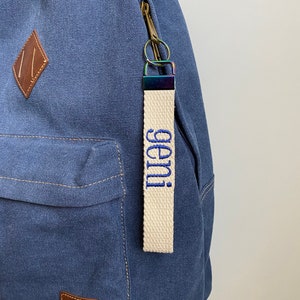 May include: A blue canvas backpack featuring a brown diamond-shaped patch and a rectangular logo patch. Attached to the zipper is a cream-colored fabric keychain strap with the word "geni" embroidered in blue thread and a metallic clasp.