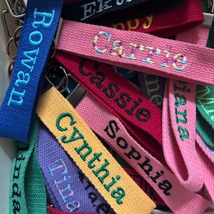 May include: A collection of fabric keychains in various colors including blue, pink, yellow, purple, green, and red. Each keychain features embroidered names such as Rowan, Ekta, Carrie, Cassie, Sophia, Cynthia, Tina, Linda, and Erin.