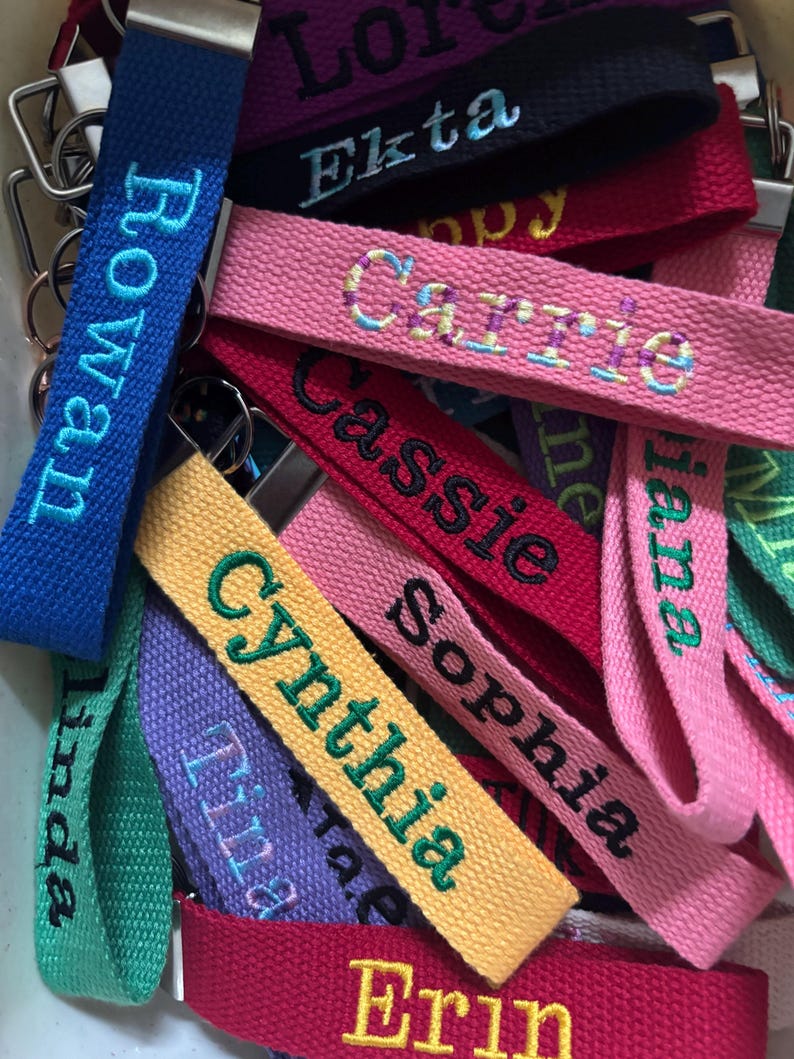 May include: A collection of fabric keychains in various colors including blue, pink, yellow, purple, green, and red. Each keychain features embroidered names such as Rowan, Ekta, Carrie, Cassie, Sophia, Cynthia, Tina, Linda, and Erin.