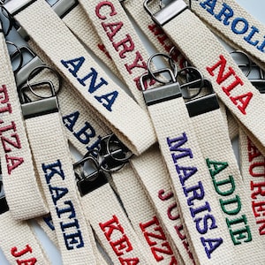 May include: A collection of off-white fabric keychains featuring embroidered names in various colors including red, blue, purple, and green. Each keychain is attached to a dark metal hardware clasp and a circular metal key ring.