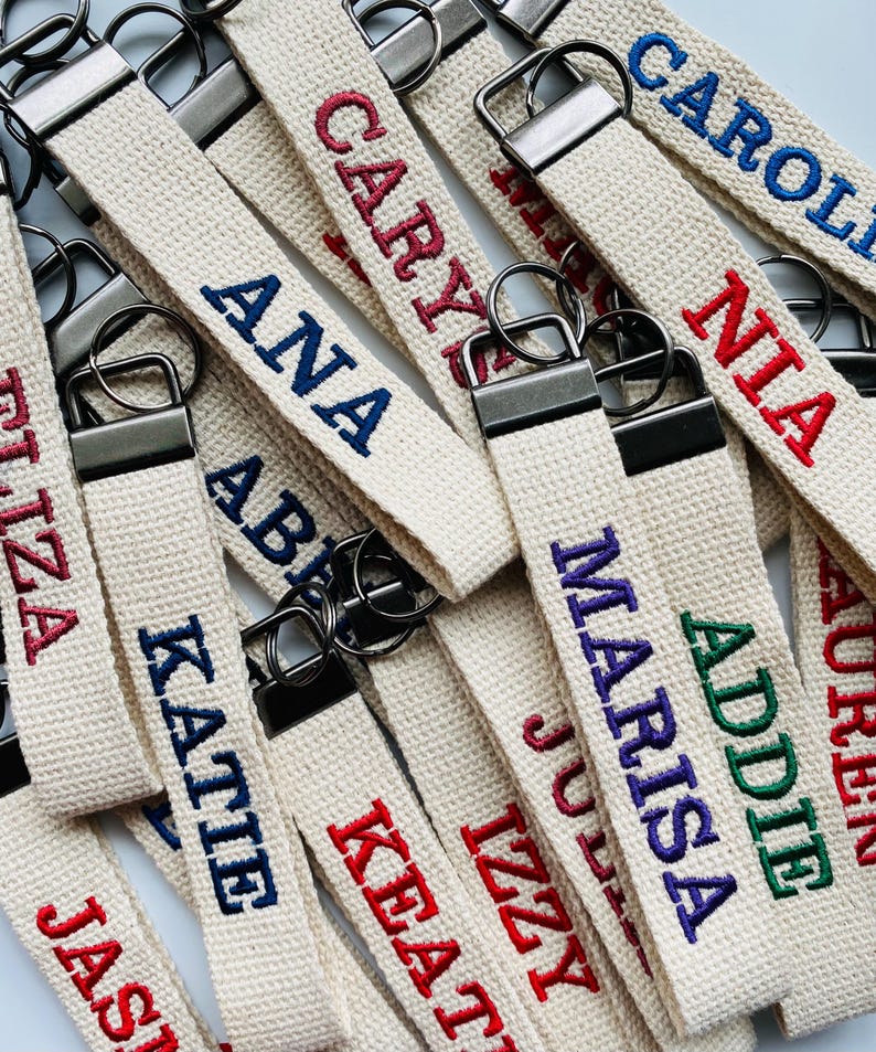 May include: A collection of off-white fabric keychains featuring embroidered names in various colors including red, blue, purple, and green. Each keychain is attached to a dark metal hardware clasp and a circular metal key ring.