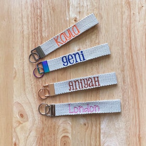 May include: Four fabric wristlet keychains in a light beige woven texture. Each features a metal clasp and ring in different finishes: gunmetal, iridescent, rose gold, and silver. Embroidered names include Kayla in orange, geni in blue, aniyah in brown, and London in pink.