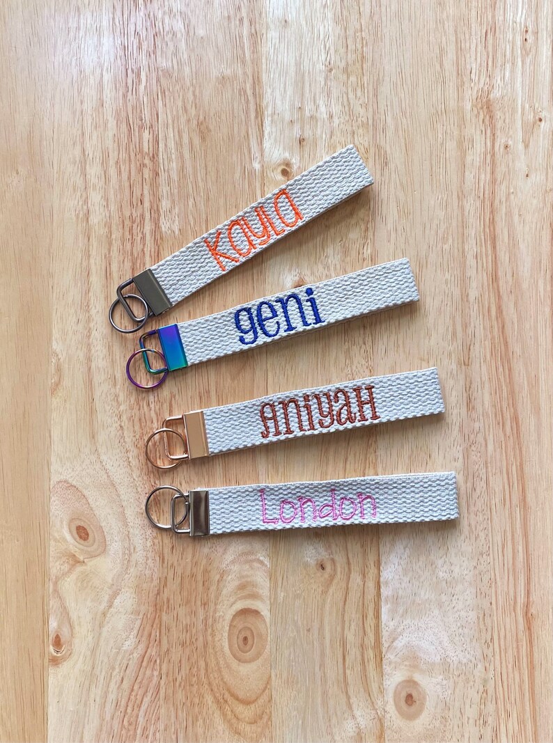 May include: Four fabric wristlet keychains in a light beige woven texture. Each features a metal clasp and ring in different finishes: gunmetal, iridescent, rose gold, and silver. Embroidered names include Kayla in orange, geni in blue, aniyah in brown, and London in pink.