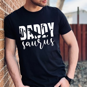 May include: A black t-shirt with a white graphic that says "Daddy-saurus" with dinosaur silhouettes.