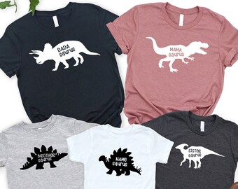 May include: Set of five t-shirts featuring dinosaur silhouettes and the text "Dada Saurus", "Mama Saurus", "Brother Saurus", "Sister Saurus", and "Name Saurus". The shirts are in various colors, including black, gray, white, and pink.