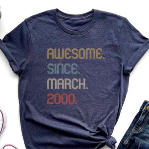 May include: A navy blue t-shirt with the text "Awesome. Since. March. 2000." in a retro font with colorful stripes.
