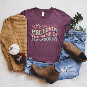 May include: A maroon t-shirt with the text "So casually cruel in the name of being honest" in a retro font with colorful flowers and hearts.