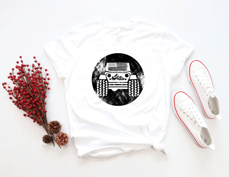 May include: White t-shirt with a black and white graphic of a Jeep with an American flag on the front. The Jeep is surrounded by a distressed circle.