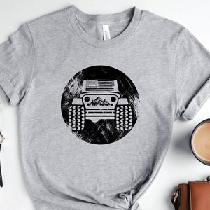 May include: Gray t-shirt with a black and white graphic of a jeep with an American flag on the hood. The jeep is in a circle with a distressed background.