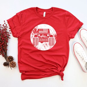 May include: Red t-shirt with a white distressed circle graphic of a jeep with an American flag on the hood and mountains in the background.