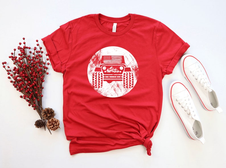May include: Red t-shirt with a white distressed circle graphic of a jeep with an American flag on the hood and mountains in the background.
