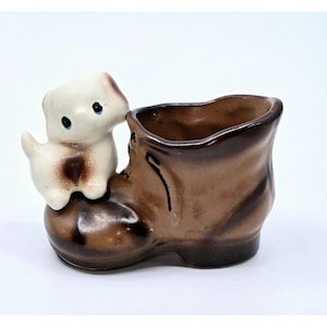 May include: A brown ceramic boot planter with a white ceramic puppy figurine sitting on top. The boot has a brown glaze and the puppy has brown spots.