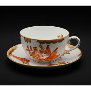 May include: A white porcelain teacup and saucer set featuring a delicate hand-painted pattern of orange and brown botanical floral motifs. The edges of both the cup and the saucer are finished with a thin gold trim, and the handle is curved.