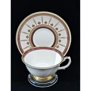 Puede incluir: Una taza de té y un platillo blancos con un borde dorado y un patrón geométrico rojo y negro. El platillo tiene un diseño dorado y negro con un borde rojo.