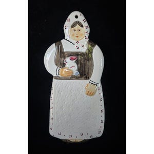 Puede incluir: Adorno de pared de cerámica con forma de mujer vestida con traje tradicional, sosteniendo un conejo blanco y rosa. La figura lleva un pañuelo blanco y un delantal con lunares. Los colores principales son blanco, marrón y rosa.