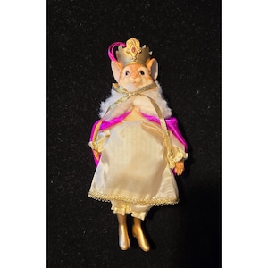Puede incluir: Figura de ratón vestido de realeza. El ratón lleva una corona dorada, una capa blanca y rosa, y un vestido color crema con adornos dorados. Los zapatos del ratón son dorados.