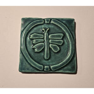May include: Square, teal-colored ceramic tile featuring a raised butterfly design within a circular frame. The butterfly has detailed wings and body. The tile has a textured surface and is likely handmade.
