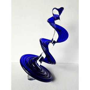 May include: A striking, abstract glass sculpture. The piece features a clear, central core spiraling upwards, wrapped by vibrant, deep blue glass ribbons. The base is a circular form, with the sculpture's height creating a dynamic, eye-catching display.