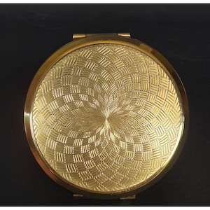 May include: A round, gold-toned metal compact mirror featuring a textured, basket-weave pattern that radiates from the center. The surface has a polished, reflective finish with a smooth, plain gold border encircling the intricate, engraved design.