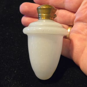 Puede incluir: Pequeño frasco de perfume de vidrio blanco con tapa de metal dorado. El frasco tiene forma redondeada con base ligeramente bulbosa y cuello cilíndrico. La tapa es ornamentada, con diseño detallado.
