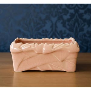 May include: A rectangular ceramic planter in a soft pastel pink hue, featuring a glossy finish with a visible crackle glaze texture. The rim is adorned with a sculpted, leaf-like pattern that wraps around the top edge of the vessel.
