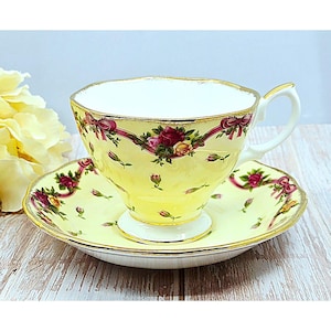 Può includere: Un set di tazza e piattino da tè giallo con un motivo floreale. La tazza ha un bordo e un manico dorati, e il piattino ha un motivo floreale abbinato. Il set è decorato con rose rosse e nastri rosa.