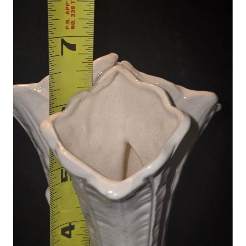 May include: A white ceramic vase with a fluted, petal-like design. A yellow measuring tape shows the vase is approximately 7 inches tall. The vase has a glossy finish and is set against a dark background.