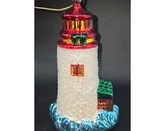 Könnte beinhalten: Weihnachtsbaumschmuck in Form eines Leuchtturms. Er hat eine rote Spitze, einen weißen, glitzernden Körper und ein kleines grünes Dach. Der Schmuck steht auf einer blauen, wellenförmigen Basis. Ein goldener Faden ist zum Aufhängen befestigt.
