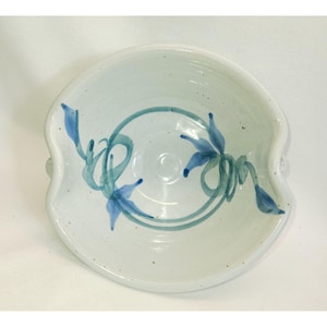 May include: A white ceramic bowl with a blue and green floral design. The bowl has a wavy rim and two small handles on the sides.