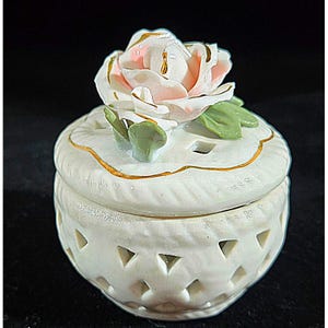 Könnte beinhalten: Weiße Keramik-Schmuckdose mit einem zarten Rosenmuster. Der Deckel zeigt eine dreidimensionale, erhabene Rose mit rosa und weißen Blütenblättern und grünen Blättern. Die Dose hat einen goldenen Rand um den Deckel und ein spitzemusterartiges Muster auf dem Boden.