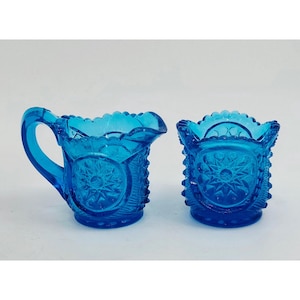 May include: Two pieces of blue glass tableware. One is a small pitcher with a handle and a scalloped rim. The other is a small, open-topped container. Both pieces have a starburst design and textured sides.