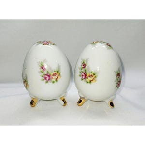 May include: A pair of white ceramic salt and pepper shakers shaped like eggs with gold feet. The shakers are decorated with a floral pattern of pink and yellow roses.