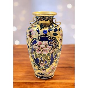 Può includere: Un vaso decorativo con bordo e base dorati, caratterizzato da un motivo floreale rosa, blu e verde. Il vaso ha uno sfondo dorato con motivi intricati e piccole maniglie. È posto su una superficie di legno.