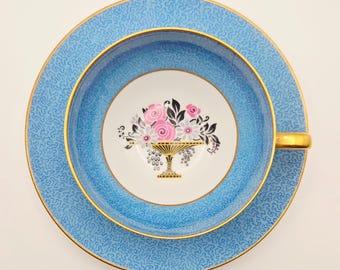 May include: A porcelain teacup and saucer set featuring a light blue background with a fine, intricate dark blue maze-like pattern. The center of the cup displays a floral arrangement in a gold-colored urn, accented with pink and white flowers.