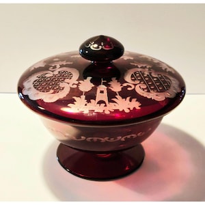 May include: A ruby red glass candy dish with a lid. The dish features etched white designs of a lighthouse, palm trees, and ornate patterns. The lid has a small, dark knob.